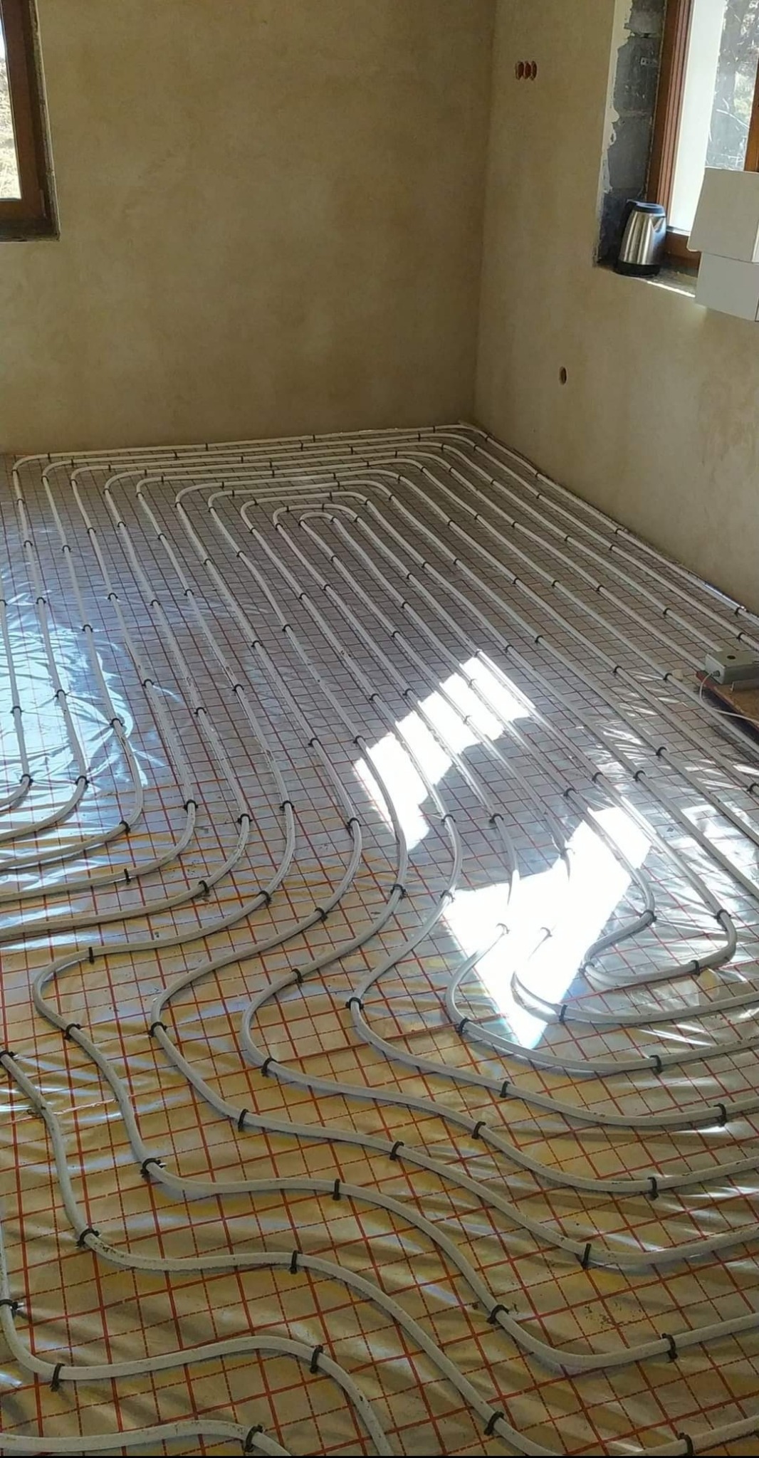 Białe rury systemu ogrzewania podłogowego rozłożone na folii z siatką w pomieszczeniu z surowymi ścianami, widoczne okno i czajnik elektryczny.