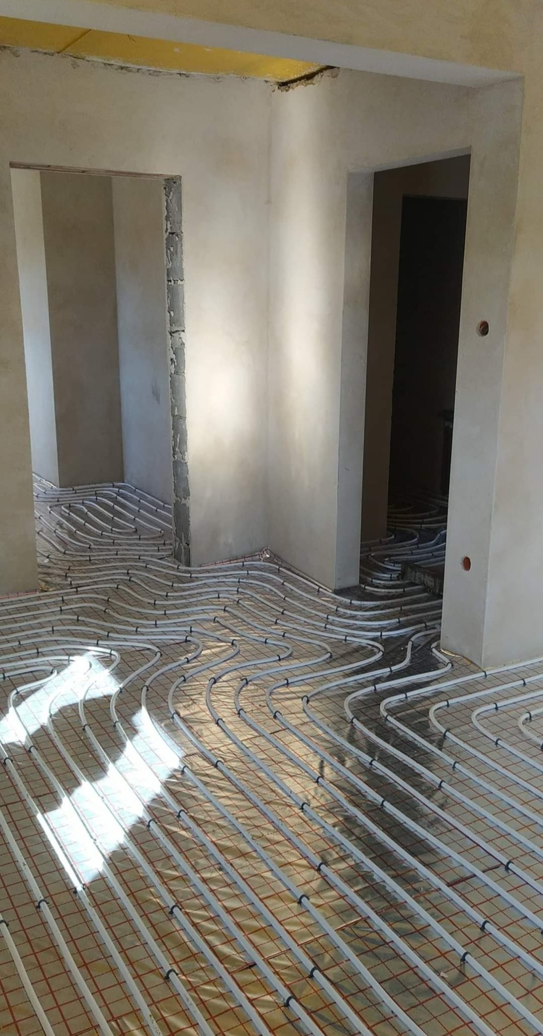 Białe rury ogrzewania podłogowego ułożone na folii w surowym wnętrzu z widocznymi otworami drzwiowymi w stanie deweloperskim.