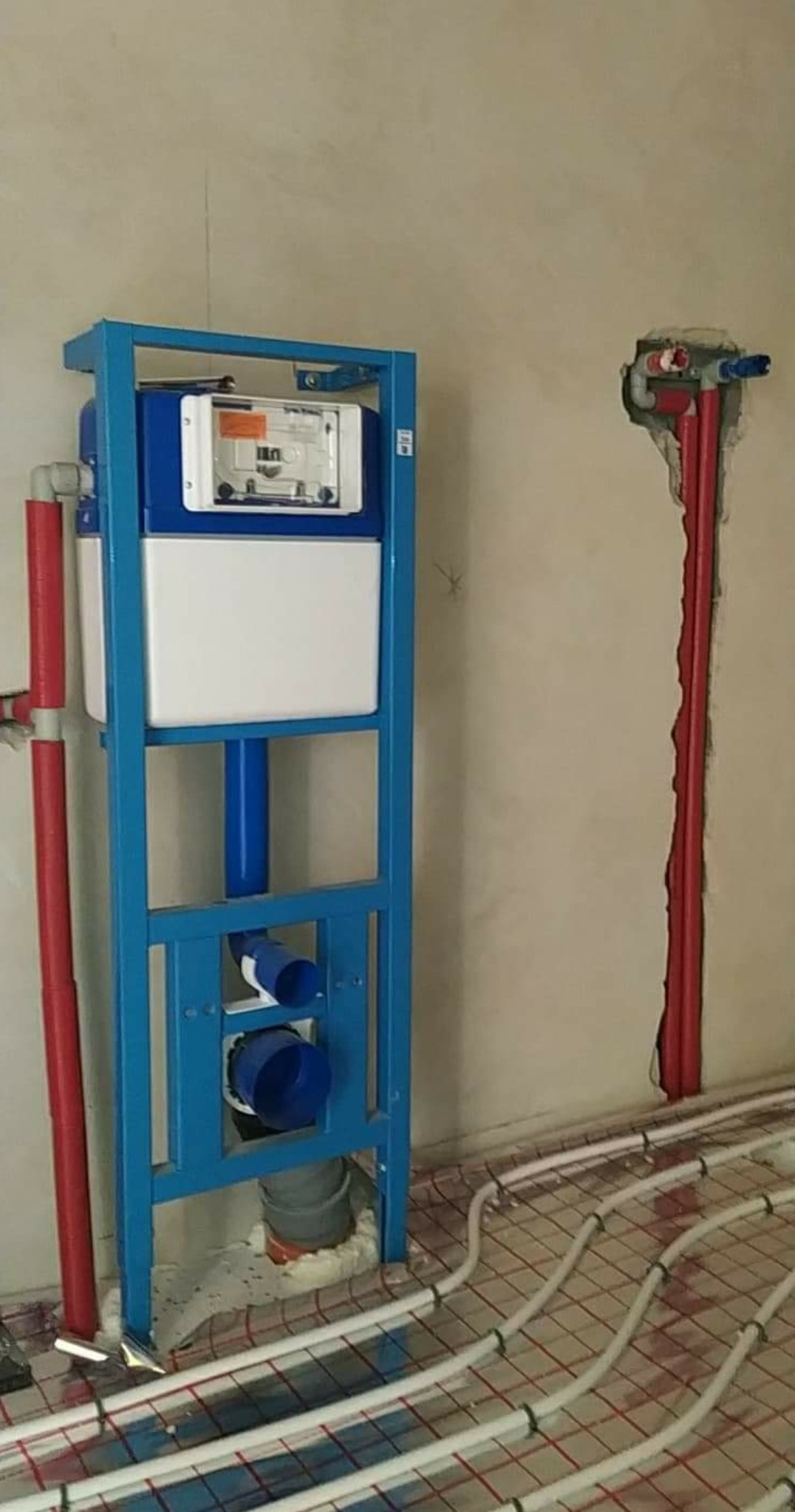 Stelaż podtynkowy WC w trakcie montażu, widoczne rury doprowadzające wodę i odpływ, instalacja ogrzewania podłogowego na podłodze, czerwone rury w ścianie.