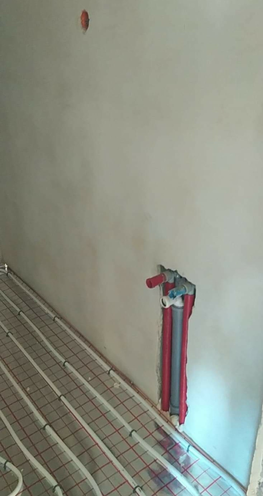 Rury instalacji wodnej wystające ze ściany nad rozłożoną matą grzewczą na podłodze w surowym stanie.