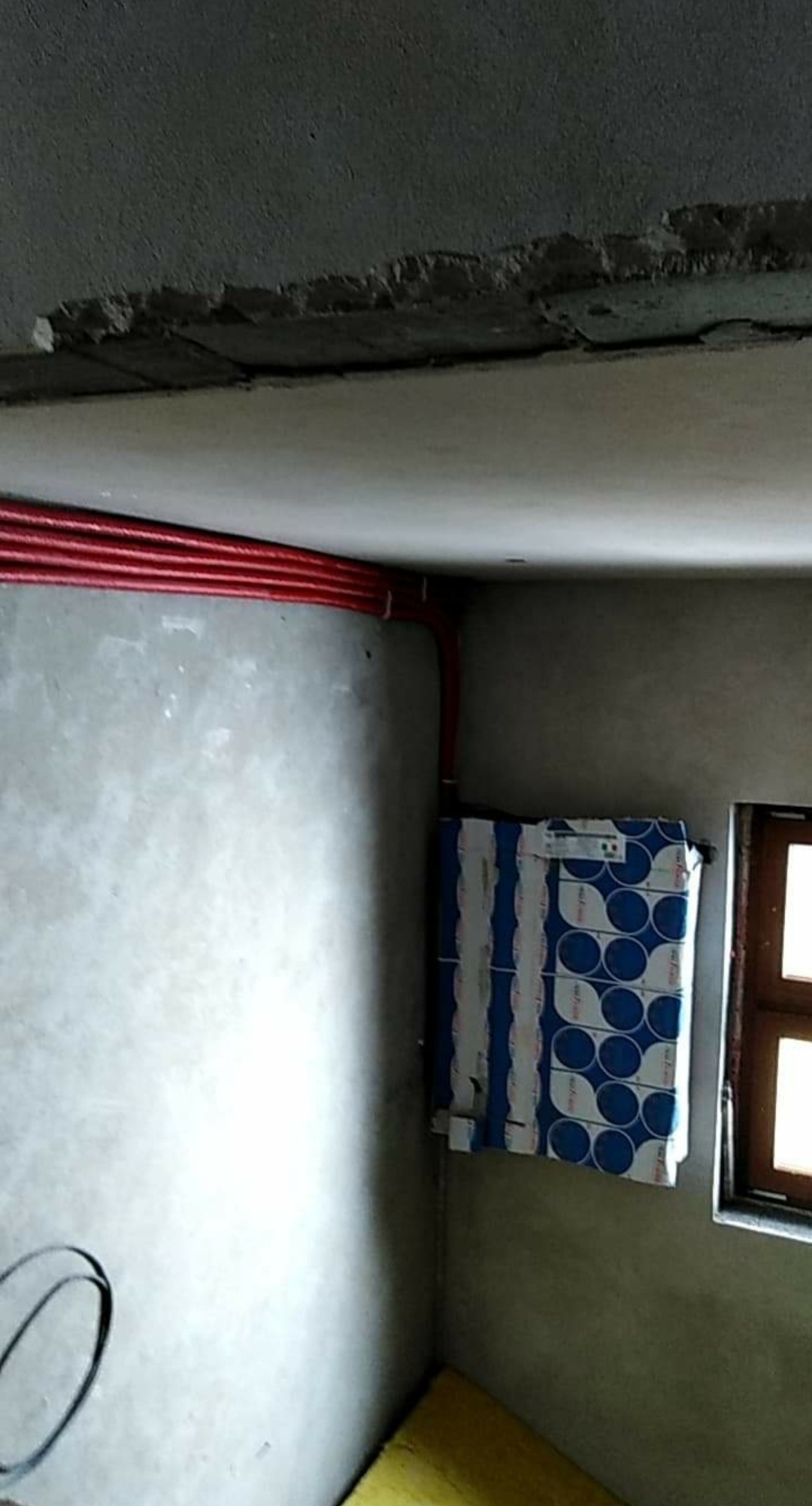 Wewnętrzny narożnik pomieszczenia w trakcie remontu; widoczne czerwone rury instalacji grzewczej poprowadzone pod sufitem, niezabezpieczony sufit z ubytkami, okno z drewnianą ramą oraz stos...