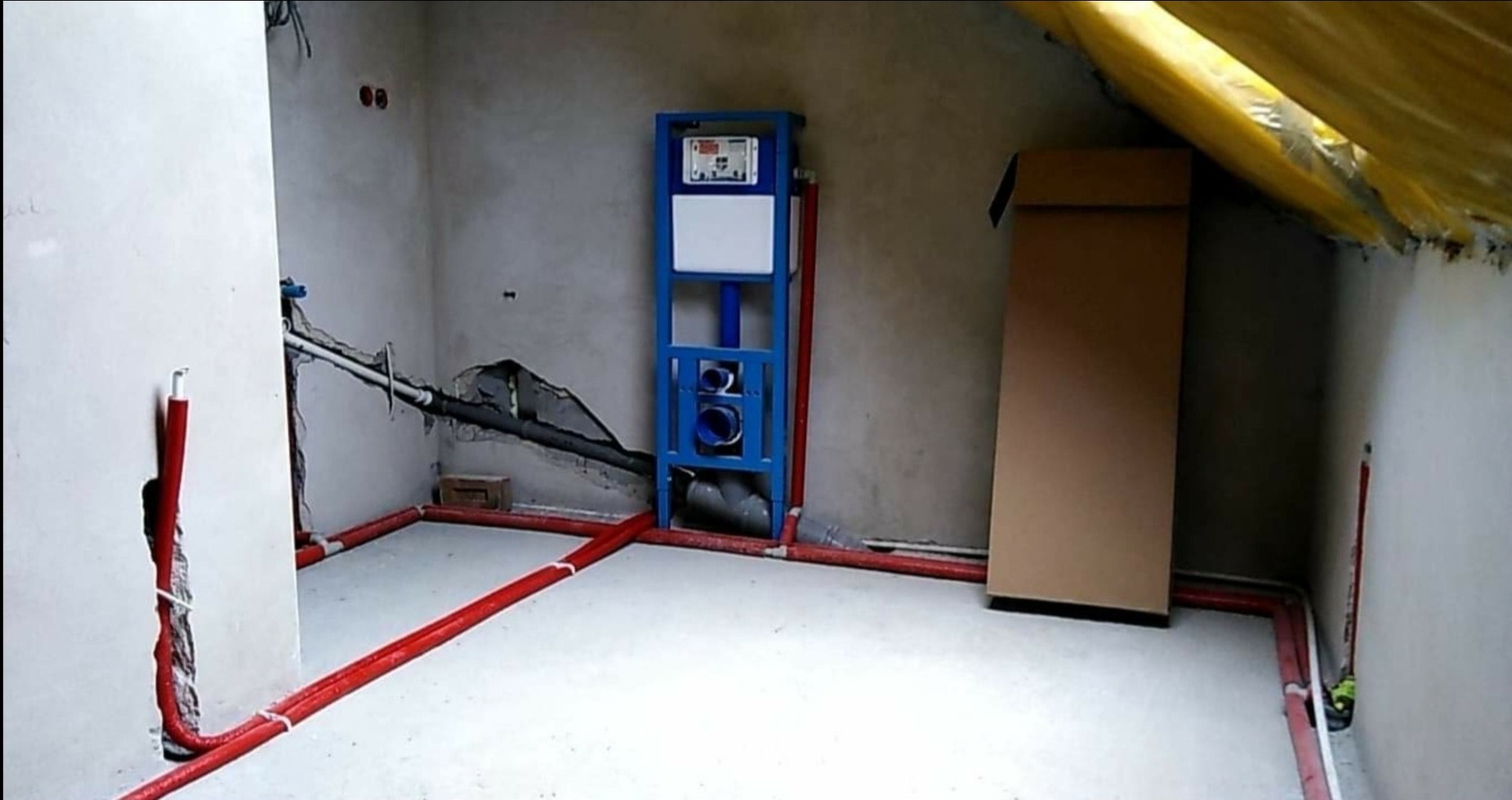 Surowy stan łazienki na poddaszu z częściowo rozprowadzoną instalacją hydrauliczną w kolorze czerwonym, widoczny stelaż podtynkowy WC w kolorze niebieskim, oraz przygotowane podejścia kanalizacyjne.