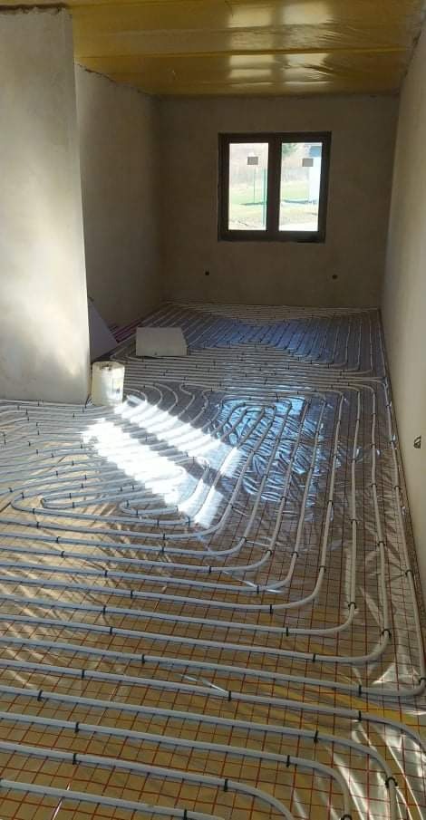 Montaż ogrzewania podłogowego w surowym pomieszczeniu, rury rozprowadzone na siatce, widoczny fragment okna i sufit z folią.