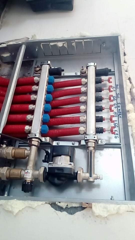 Rozdzielacz instalacji ogrzewania podłogowego z czerwonymi rurami w metalowej szafce, widoczne zawory, pompa obiegowa i izolacja piankowa wokół szafki.