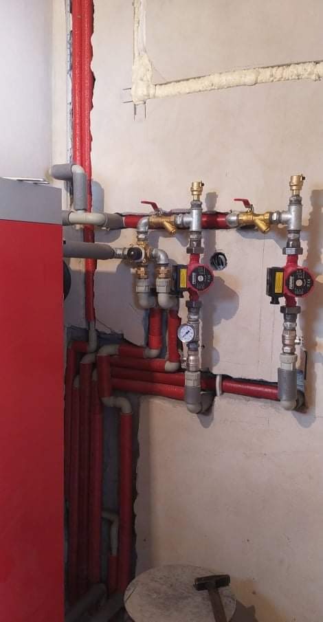 Rozbudowana instalacja grzewcza z czerwonymi rurami, pompami obiegowymi i zaworami na tle surowej ściany z widocznymi śladami prac instalacyjnych.
