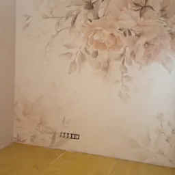 Ściana z motywem akwarelowych kwiatów w odcieniach beżu i różu, widoczne gniazdka elektryczne, podłoga zabezpieczona folią malarską, w rogu narzędzia i wiadro.
