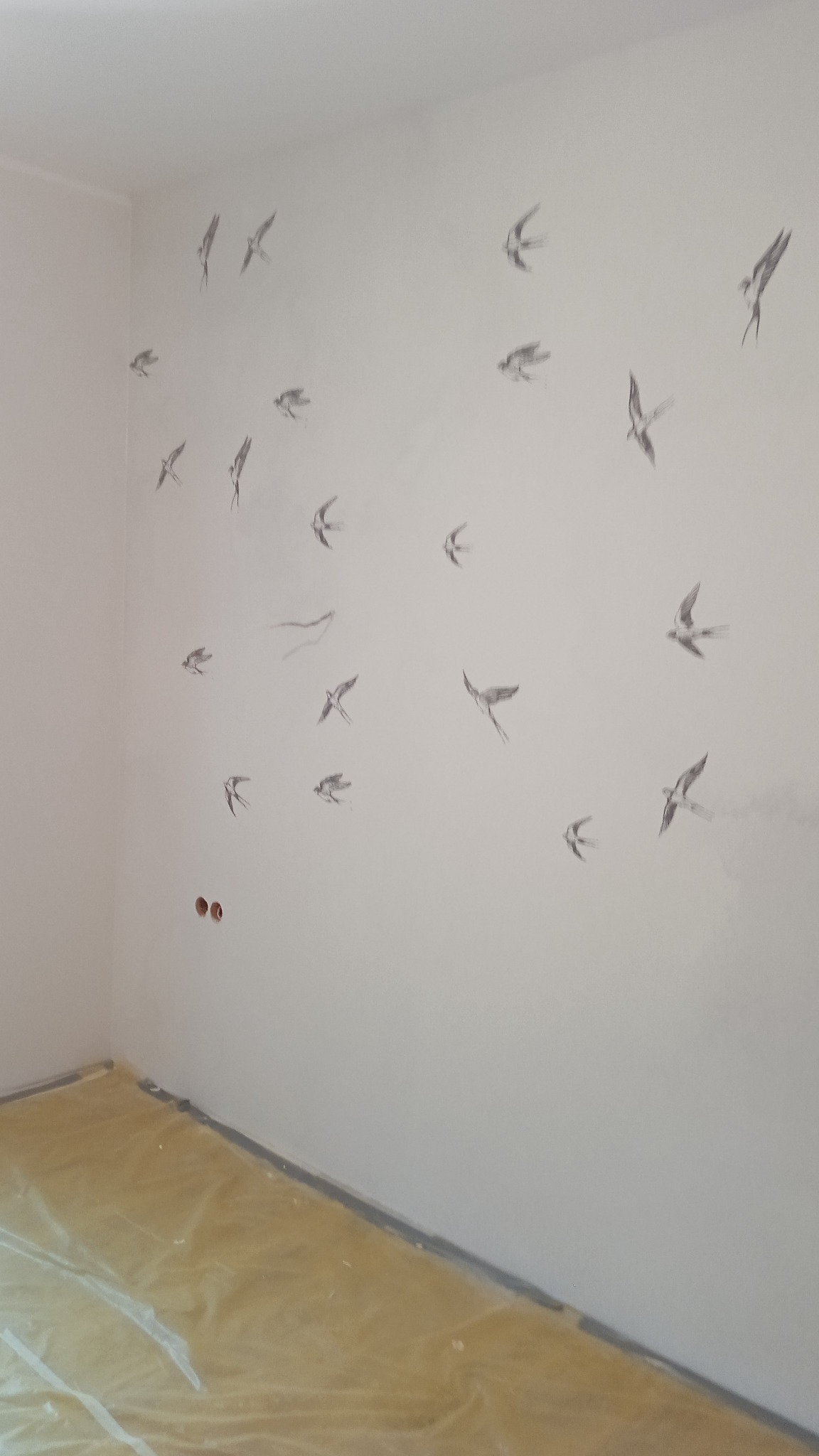 Biała ściana w pokoju z motywem ręcznie malowanych jaskółek w różnych pozach; podłoga zabezpieczona folią malarską.