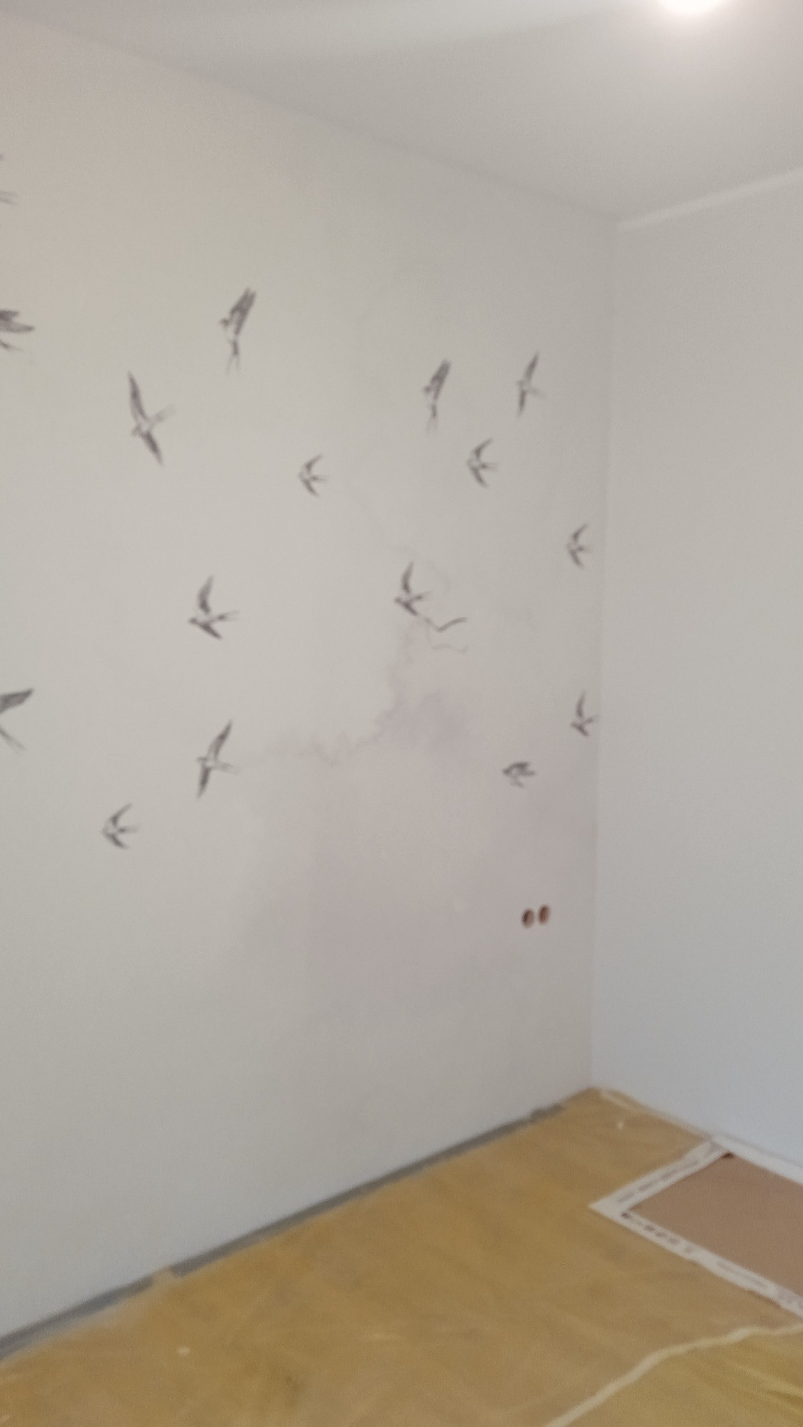 Biała ściana w rogu pokoju z motywem szarych jaskółek, widoczne ślady po wcześniejszych pracach na ścianie, podłoga zabezpieczona folią malarską.
