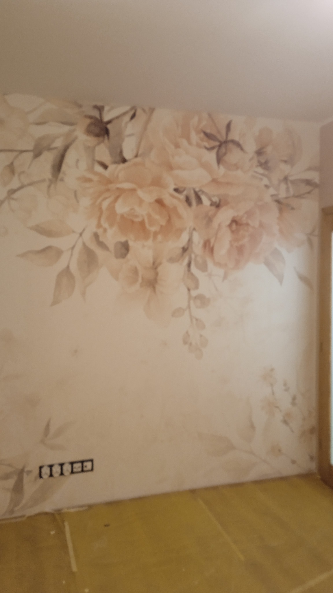Ściana w pokoju z delikatnym, rozmytym wzorem kwiatowym w odcieniach beżu i brązu, widoczne gniazdka elektryczne przy podłodze i fragment drewnianej futryny drzwi.