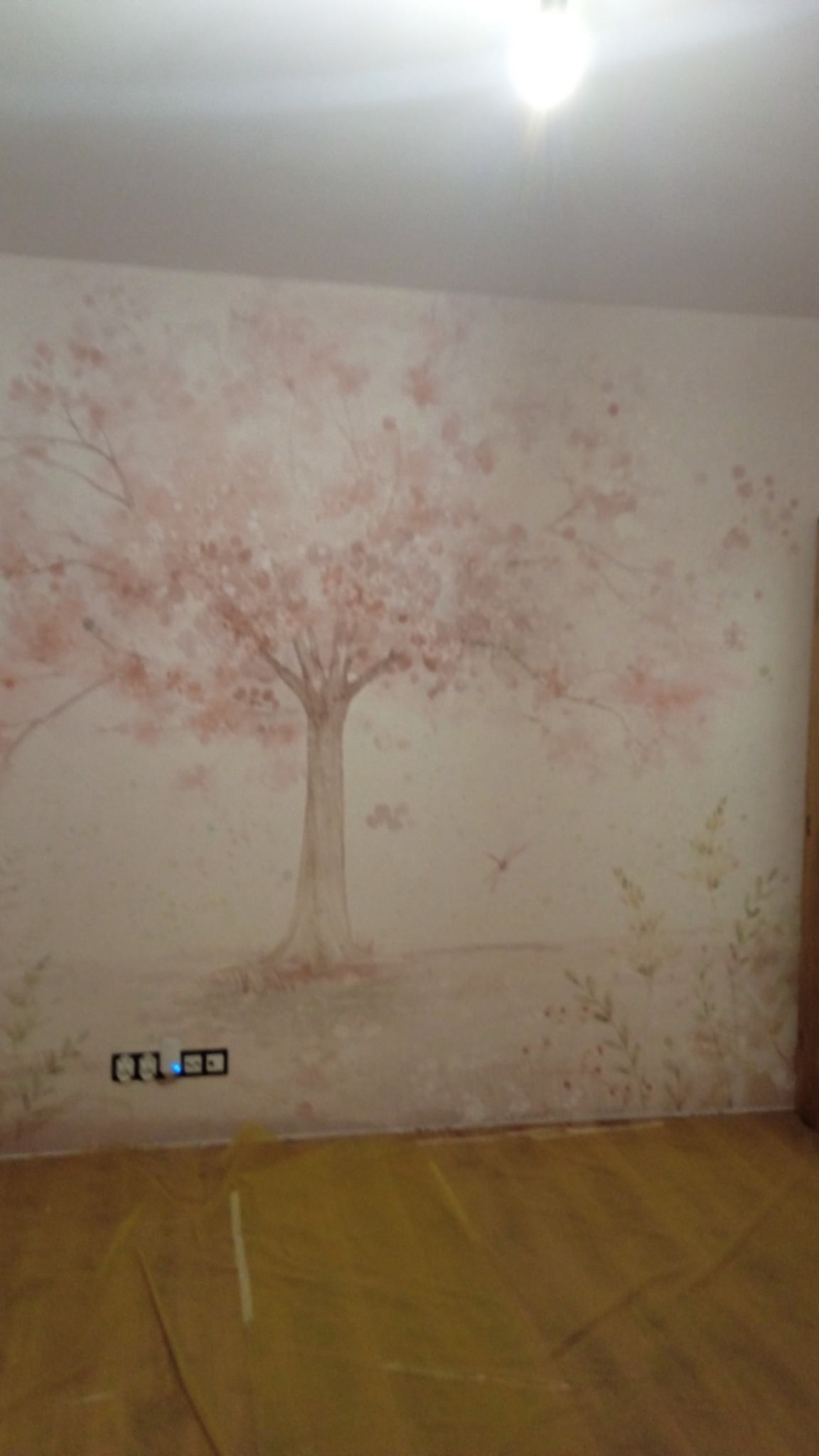 Ściana w pokoju z ręcznie malowanym motywem drzewa wiśni w odcieniach różu i szarości, zabezpieczona folią podłoga, widoczne gniazdka elektryczne.
