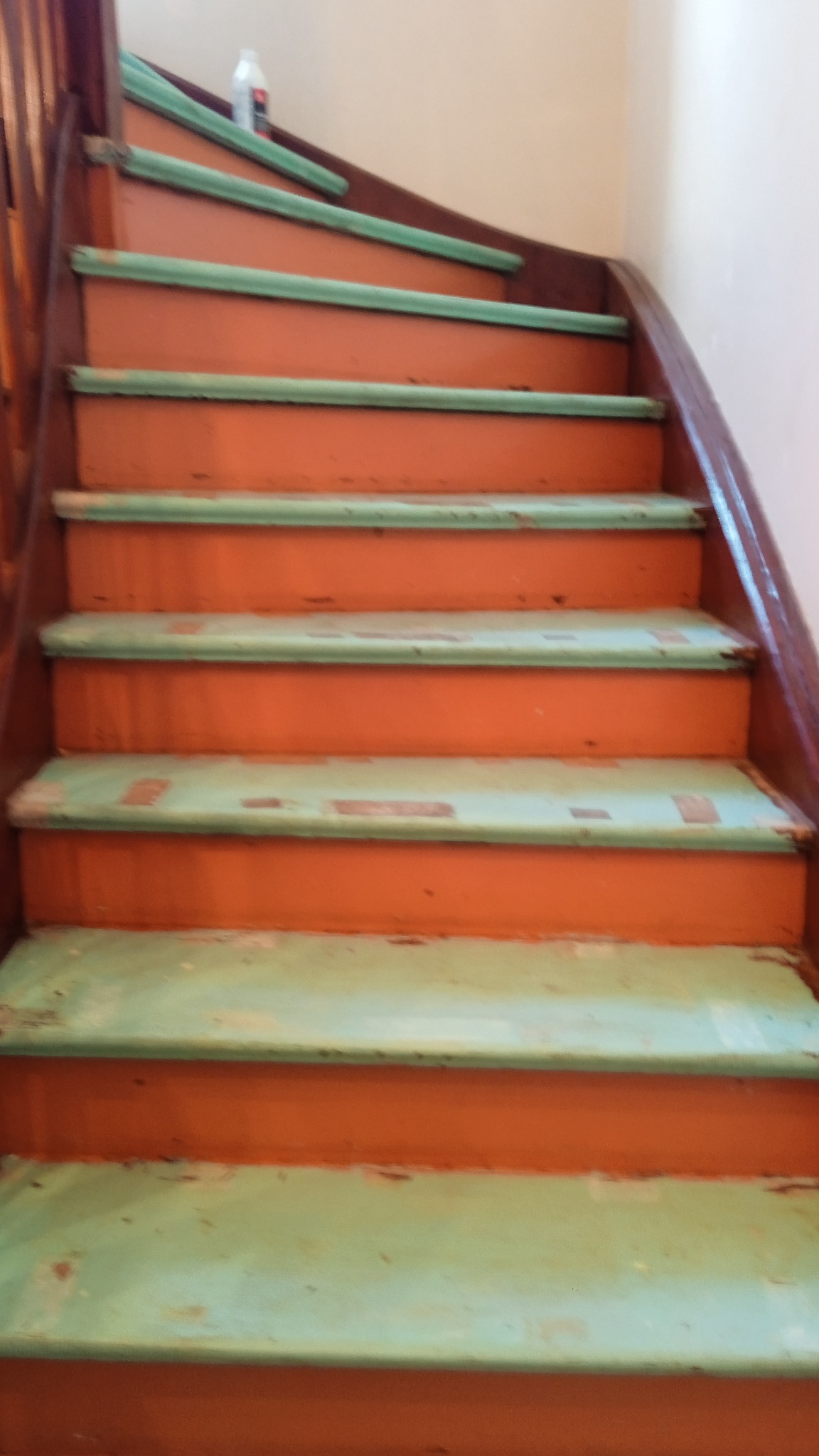 Schody z pomarańczowymi podstopnicami i zielonymi krawędziami stopni, widoczne ślady użytkowania i odchodząca farba, butelka z płynem na górnym stopniu.