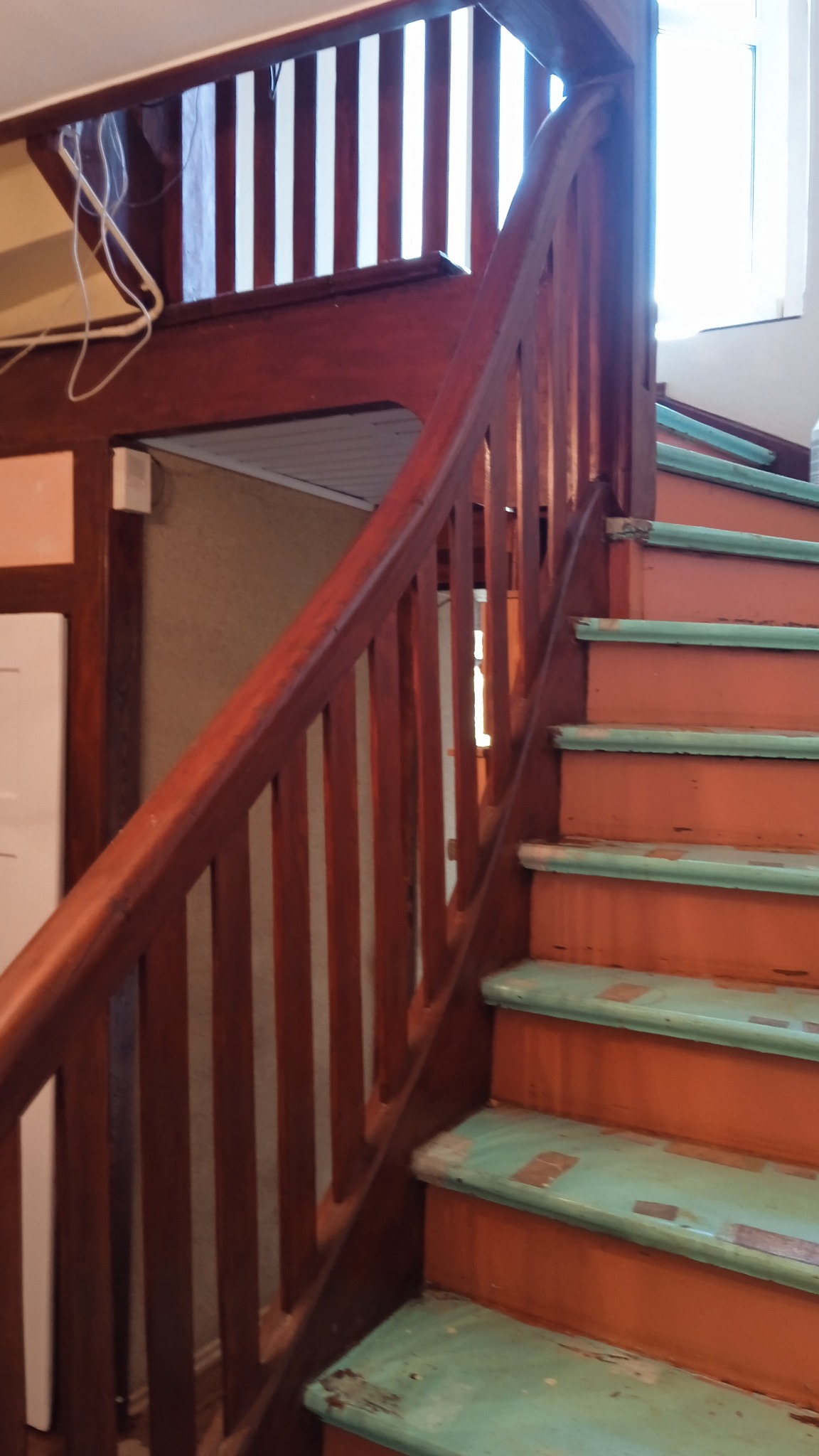 Drewniane schody z pomalowanymi na pomarańczowo stopniami i zielonymi krawędziami, widoczne ślady zużycia i odpryski farby, brązowa balustrada i poręcz, fragment wnętrza domu.