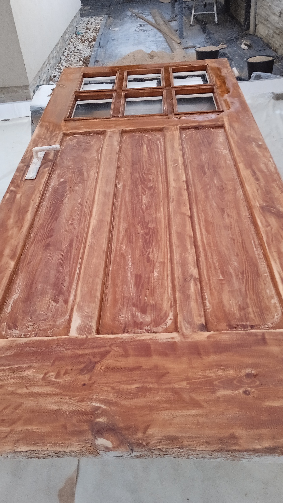 Drewniane drzwi z szybkami, świeżo polakierowane na brązowo, leżące na białym podłożu w trakcie renowacji.