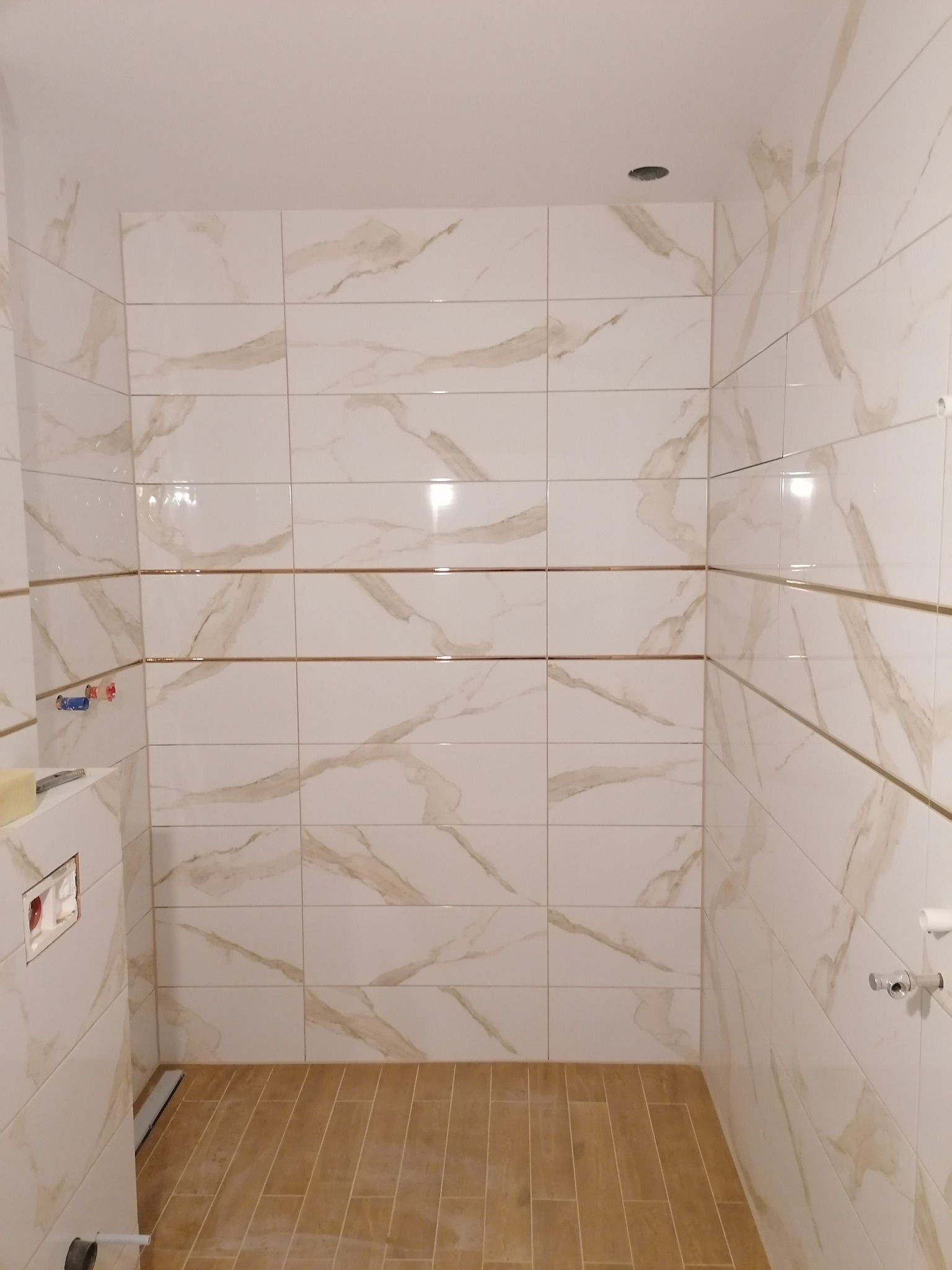 Wnętrze łazienki w trakcie remontu: białe płytki marmurowe z subtelnymi złotymi żyłkami na ścianach, drewnopodobna podłoga, widoczne elementy instalacji sanitarnej przed montażem.