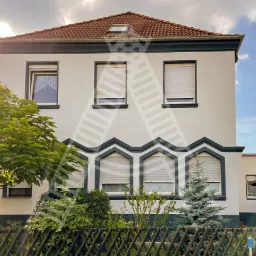 Dwukondygnacyjny dom z białymi ścianami i ciemnozielonymi akcentami, widoczny zza drewnianego płotu i zieleni, okna z żaluzjami opuszczonymi, dach z czerwonej dachówki.