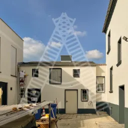 Pracownik na drabinie maluje elewację budynku w zabudowie szeregowej na tle błękitnego nieba z chmurami, widoczne pozostałości materiałów budowlanych.