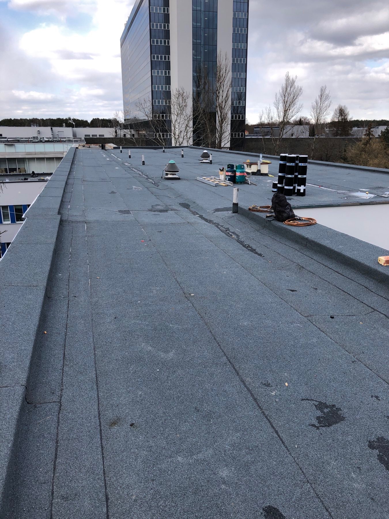 Płaski dach pokryty papą termozgrzewalną z widocznymi zgrzewami, kominkami wentylacyjnymi i narzędziami pracy dekarza, na tle budynku biurowego i pochmurnego nieba.