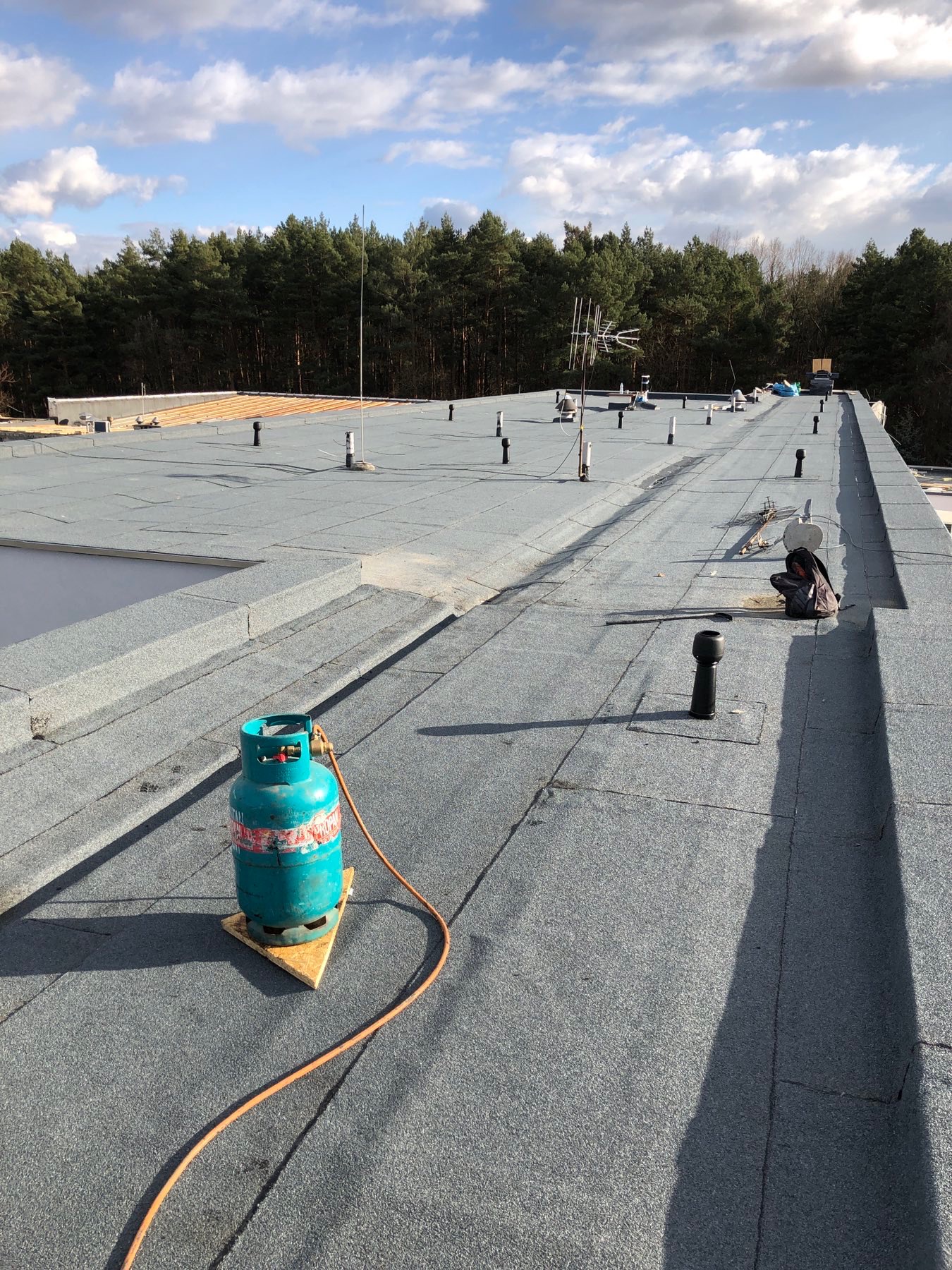 Zielona butla gazowa na drewnianym podeście, leżąca na szarym, płaskim dachu pokrytym papą termozgrzewalną. W tle widoczny las iglasty i błękitne niebo z chmurami. Na dachu kominki wentylacyjne...