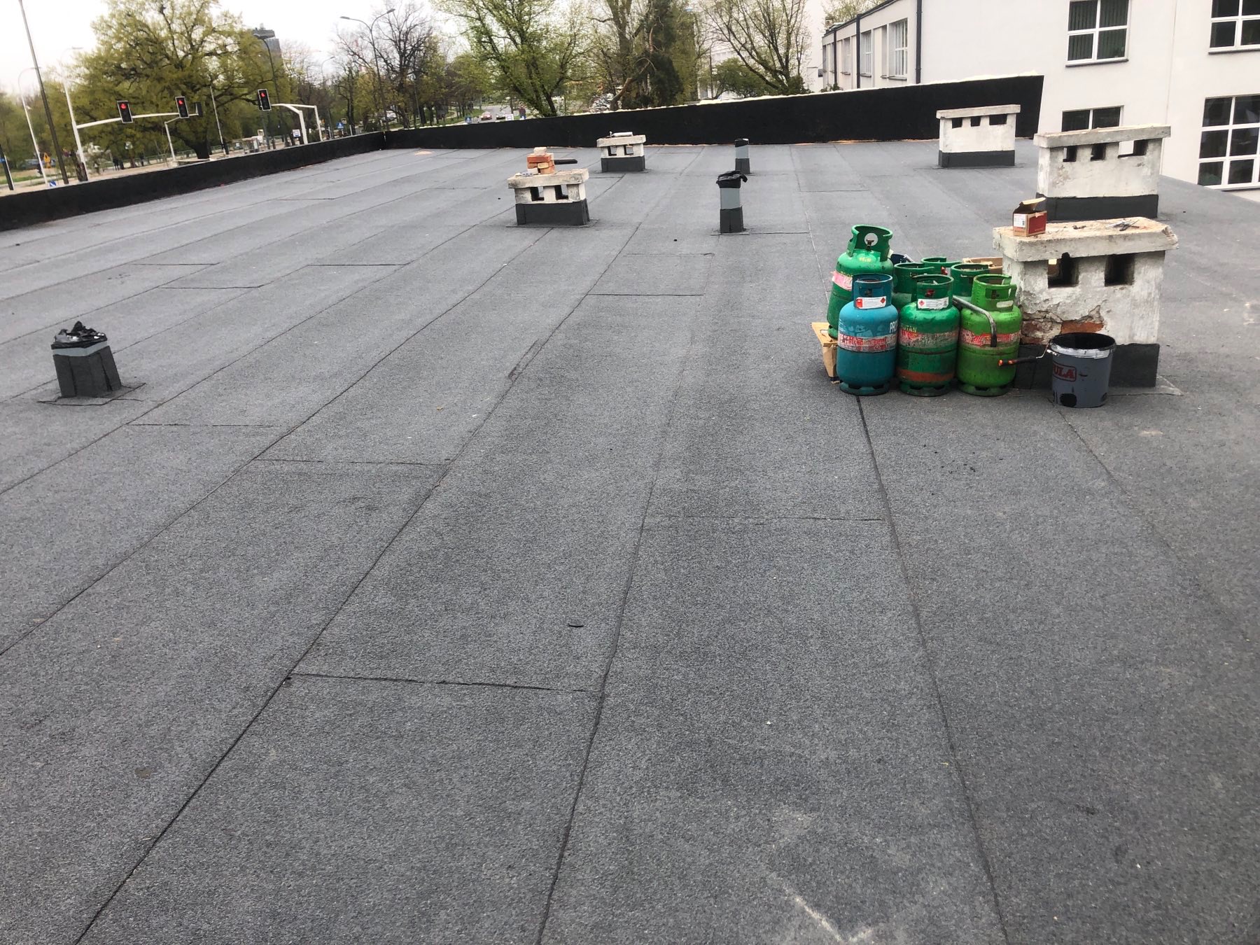 Płaski dach pokryty papą termozgrzewalną, widoczne kominy wentylacyjne i butle gazowe używane podczas prac dekarskich.