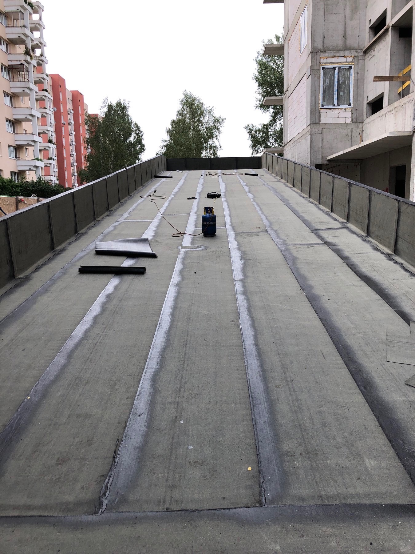 Płaski dach pokryty papą termozgrzewalną z widocznymi zgrzewami, butlą gazową i narzędziami na tle budynków mieszkalnych w trakcie budowy.