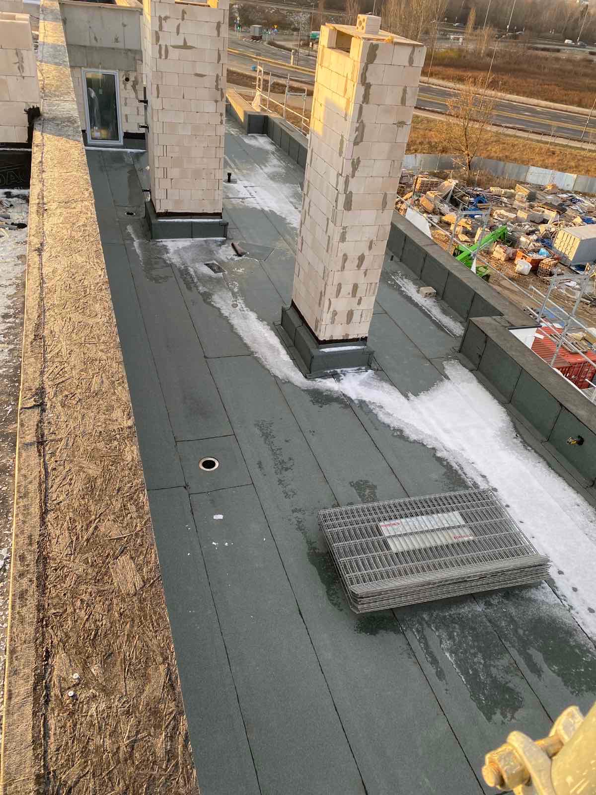 Widok z góry na płaski dach pokryty papą termozgrzewalną, z wystającymi kominami z bloczków i stos krat pomostowych, częściowo pokryty śniegiem, z widocznym fragmentem elewacji budynku i elementami...