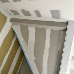 FIRMA BUDOWLANA JAKUB WISZNIEWSKI - Wnętrze w trakcie prac wykończeniowych: ściany z płyt kartonowo-gipsowych z widocznymi łączeniami szpachlowanymi, częściowo pomalowane, widoczna konstrukcja sufitu podwieszanego z profilami...