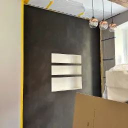 Ściana w trakcie prac wykończeniowych: czarna, fakturowana powierzchnia z wnękami, zabezpieczona taśmą malarską, widoczne rury przy podłodze, elementy oświetlenia i płyta karton-gips.