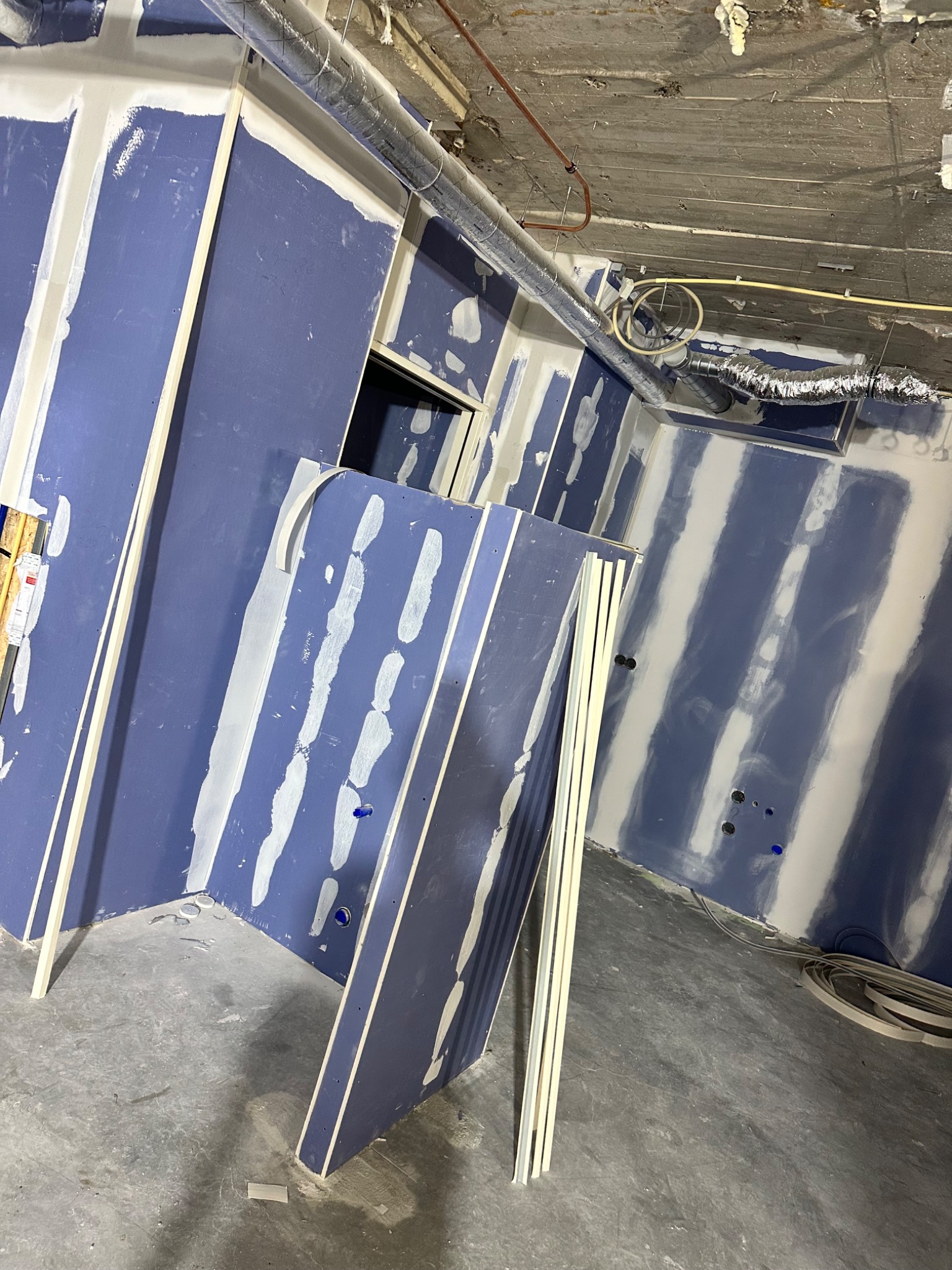 Wnętrze w trakcie prac wykończeniowych: niebieskie ściany z płyt kartonowo-gipsowych, częściowo zaszpachlowane, widoczne instalacje pod sufitem.