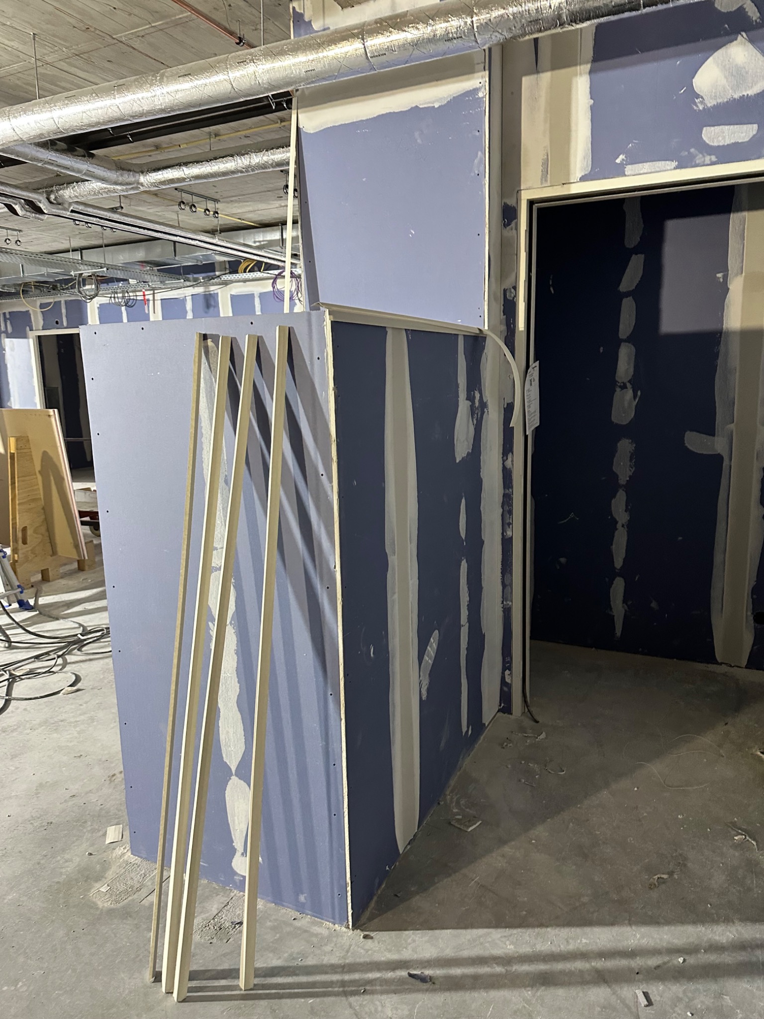 Wnętrze w trakcie prac wykończeniowych: widoczne niebieskie płyty gipsowo-kartonowe z widocznymi łączeniami, profile drewniane, instalacje pod sufitem, surowy beton na podłodze, fragment otworu...