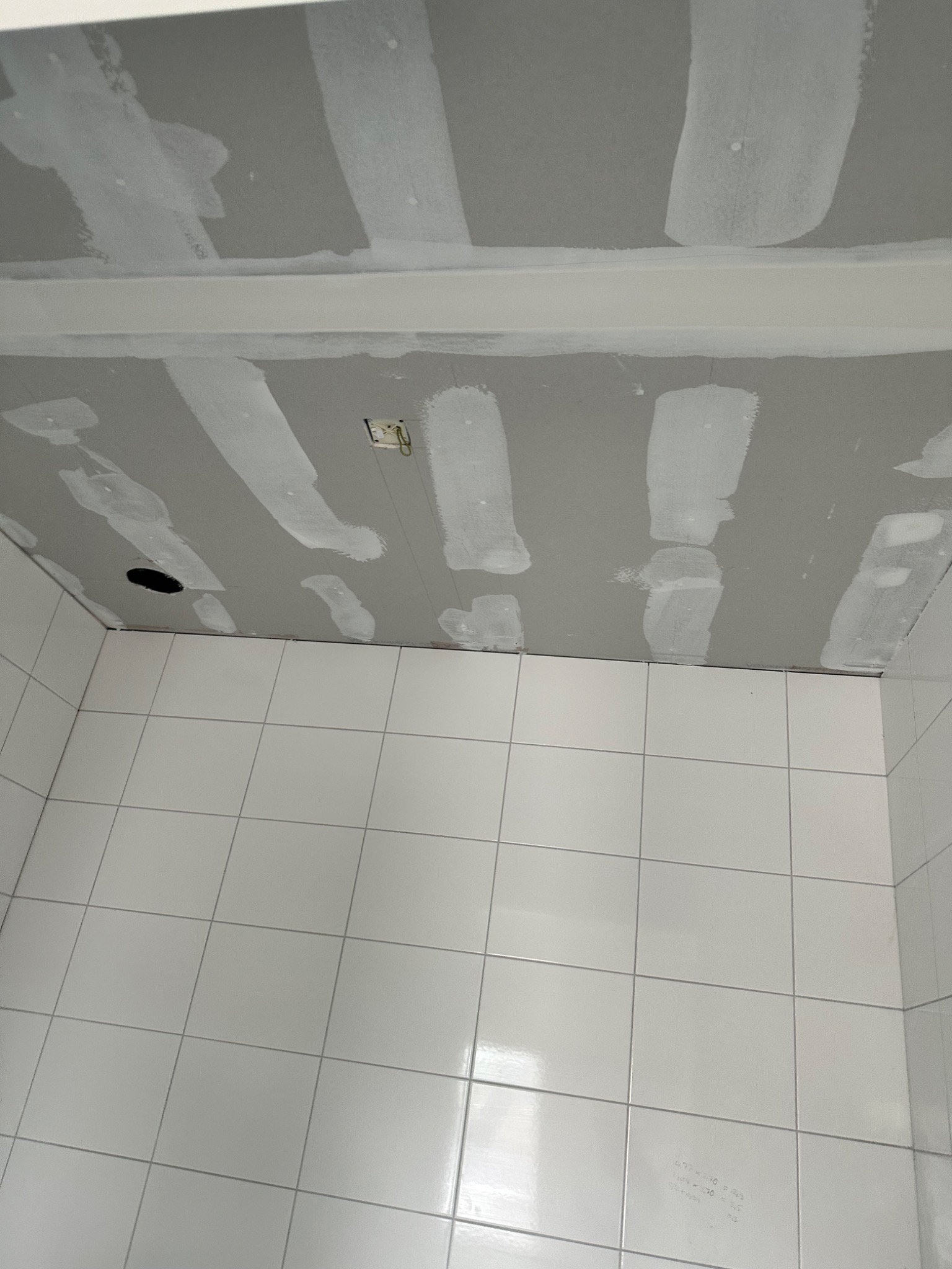 Świeżo zamontowana płyta kartonowo-gipsowa na suficie łazienki, z widocznymi łączeniami zaszpachlowanymi na biało, otwór na oświetlenie i białe płytki na ścianach.