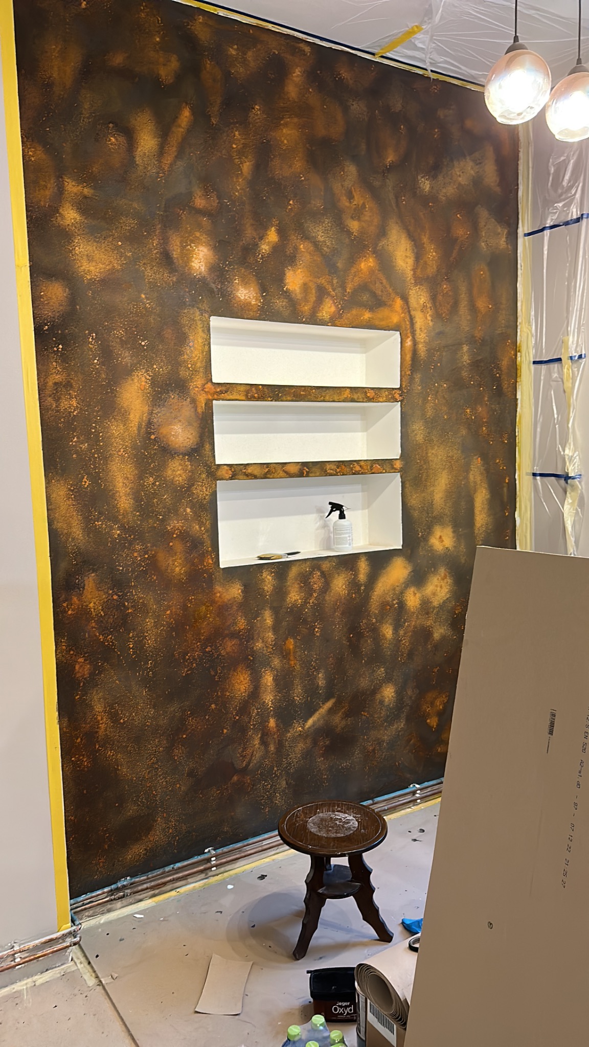Ściana z dekoracyjną, rdzawą fakturą i wnękami z białymi półkami, widoczne narzędzia i akcesoria malarskie, fragment płyty karton-gips na pierwszym planie, oświetlenie sufitowe.