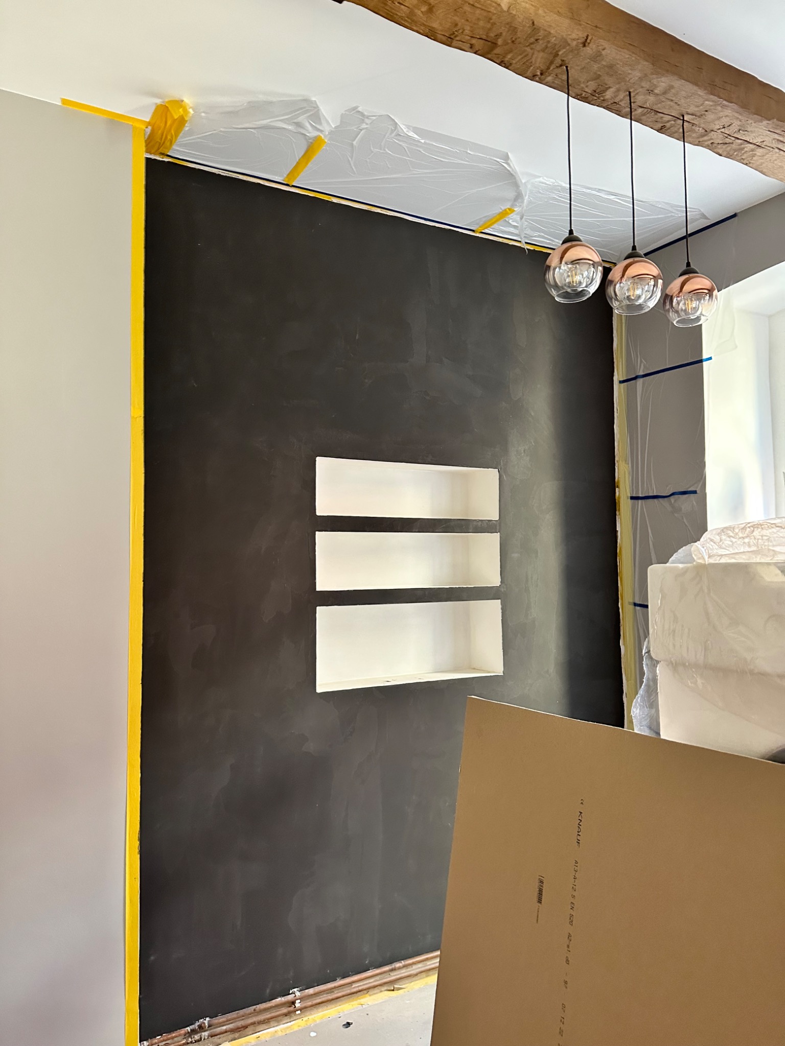 Ściana w trakcie prac wykończeniowych: czarna, fakturowana powierzchnia z wnękami, zabezpieczona taśmą malarską, widoczne rury przy podłodze, elementy oświetlenia i płyta karton-gips.
