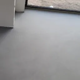 Szara, gładka posadzka w nowym budynku, widok na zewnątrz przez okno zabezpieczone folią malarską.