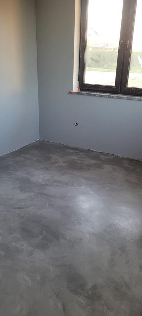 Pokój po remoncie z szarą, gładką posadzką imitującą beton i ścianami w odcieniu gołębiej szarości, widoczne okno z czarną ramą.