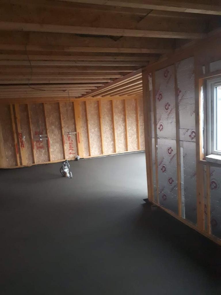Świeżo wylana, ciemnoszara posadzka w surowym wnętrzu z drewnianą konstrukcją ścian i izolacją termiczną; widoczny sprzęt do wylewania posadzek.