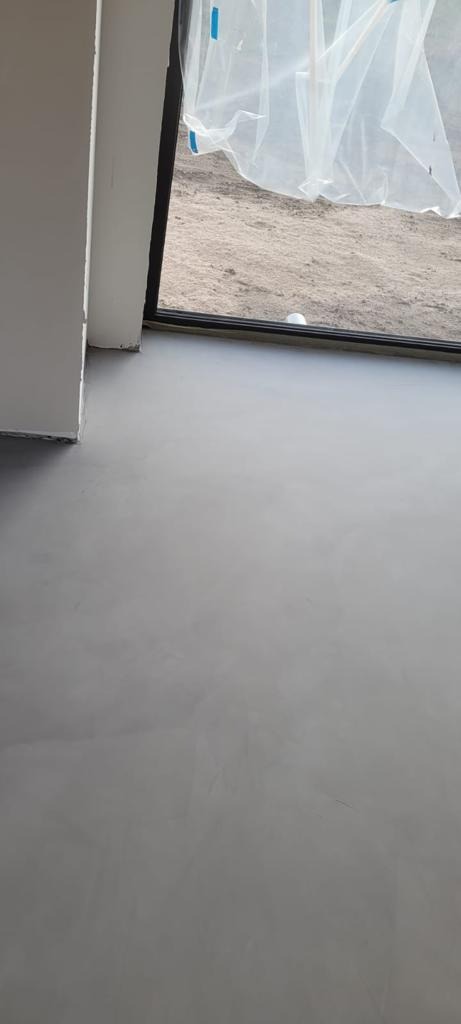 Szara, gładka posadzka w nowym budynku, widok na zewnątrz przez okno zabezpieczone folią malarską.