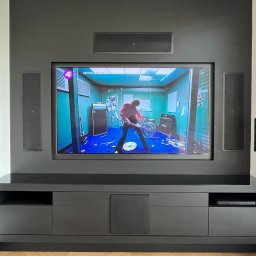 CosmoHomes Nowak & Szerfenberg Sp. z o.o. - Szara zabudowa ściany z telewizorem i głośnikami w salonie, na ekranie telewizora scena z muzykiem i perkusją, szafka RTV z szufladami i półkami.