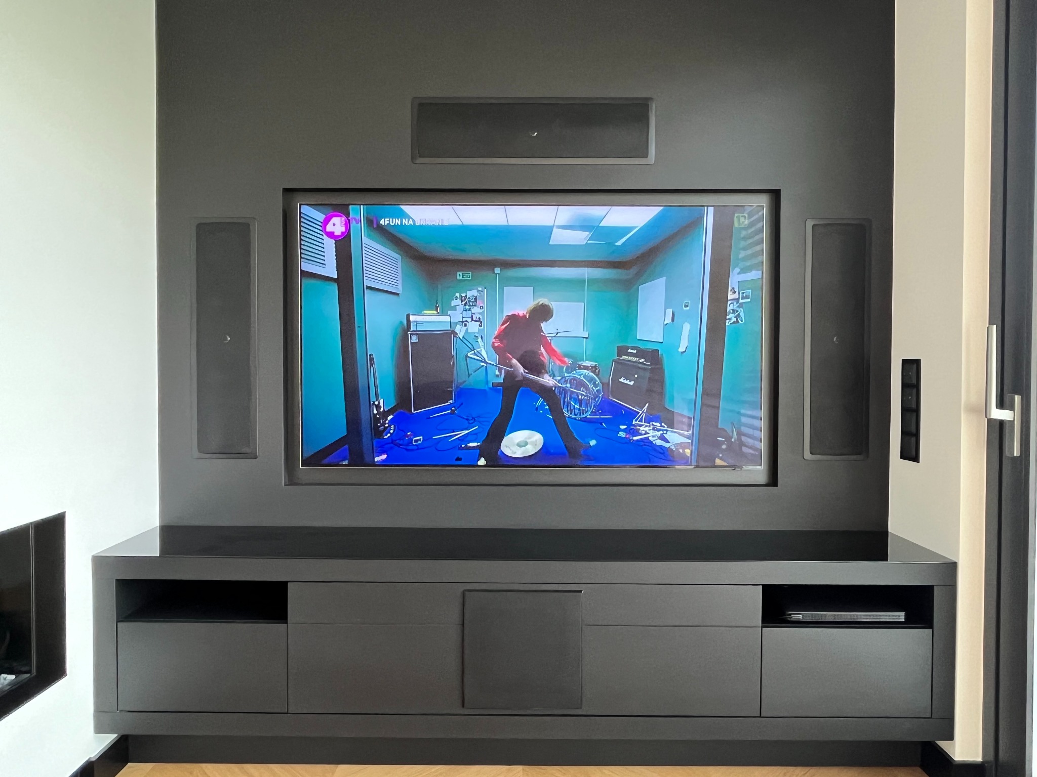 Szara zabudowa ściany z telewizorem i głośnikami w salonie, na ekranie telewizora scena z muzykiem i perkusją, szafka RTV z szufladami i półkami.