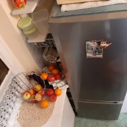 Szara lodówka z magnesami (słoń i sowa) stojąca w kuchni obok koszyka z jabłkami i pomarańczami, z widoczną zawartością półek i pojemników na drzwiach.