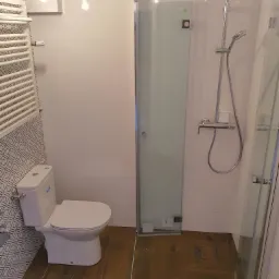 Nowoczesna łazienka po remoncie: kabina prysznicowa ze szklanymi drzwiami, toaleta kompaktowa z wzorzystą ścianą, grzejnik drabinkowy i brązowe płytki podłogowe imitujące drewno.