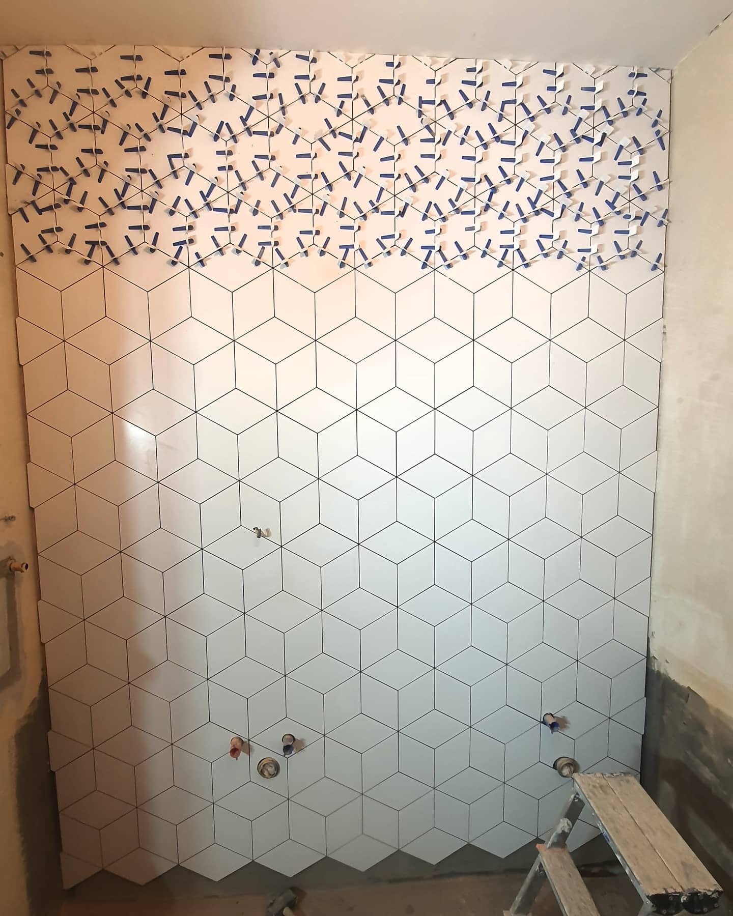 Ściana w trakcie układania białych, heksagonalnych płytek z czarnym konturem, z widocznymi niebieskimi krzyżykami dystansowymi w górnej części, surowy stan wykończenia ścian bocznych, fragment...