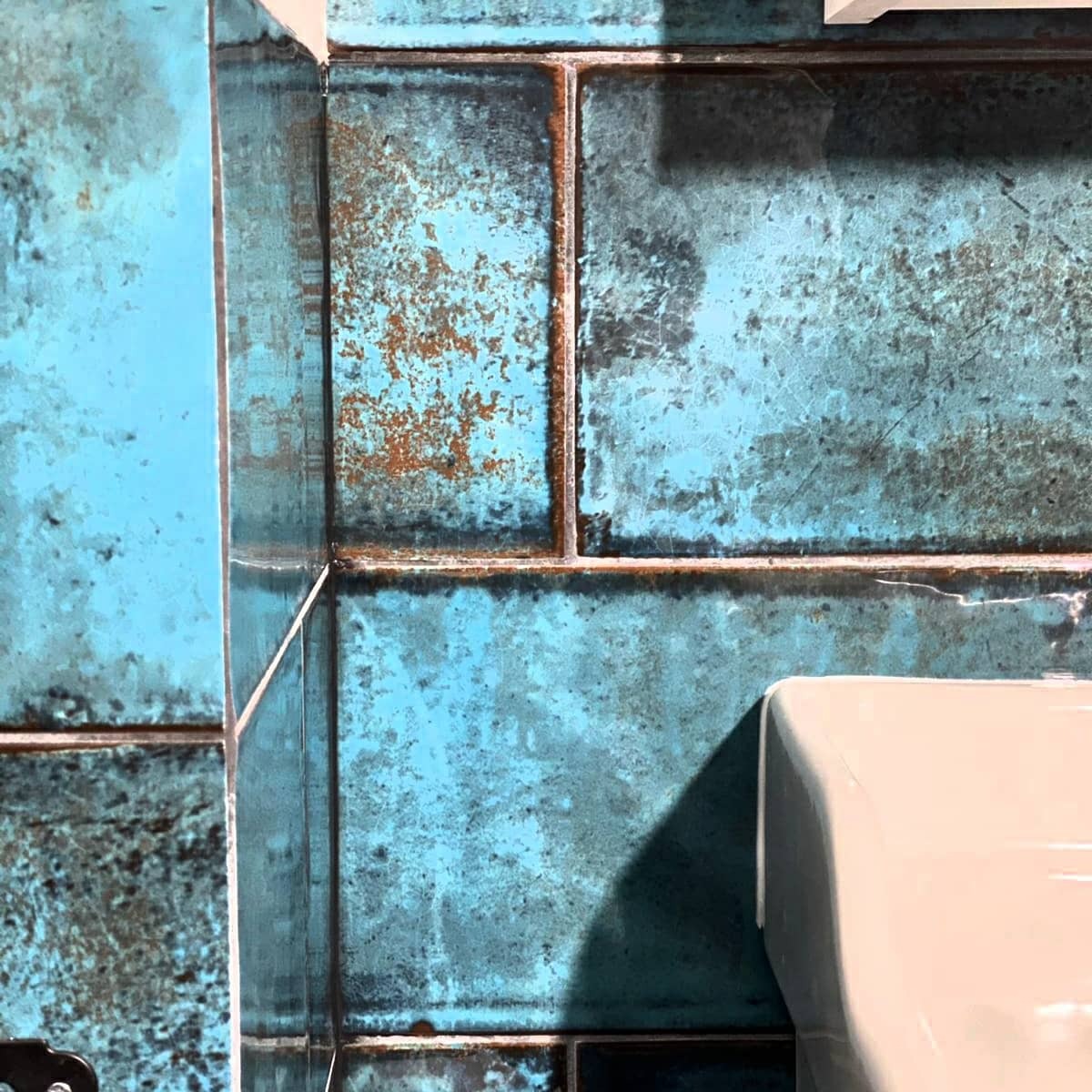 Fragment ściany wyłożonej turkusowymi płytkami z efektem patyny i rdzawymi akcentami, widoczny narożnik i fragment białej umywalki.