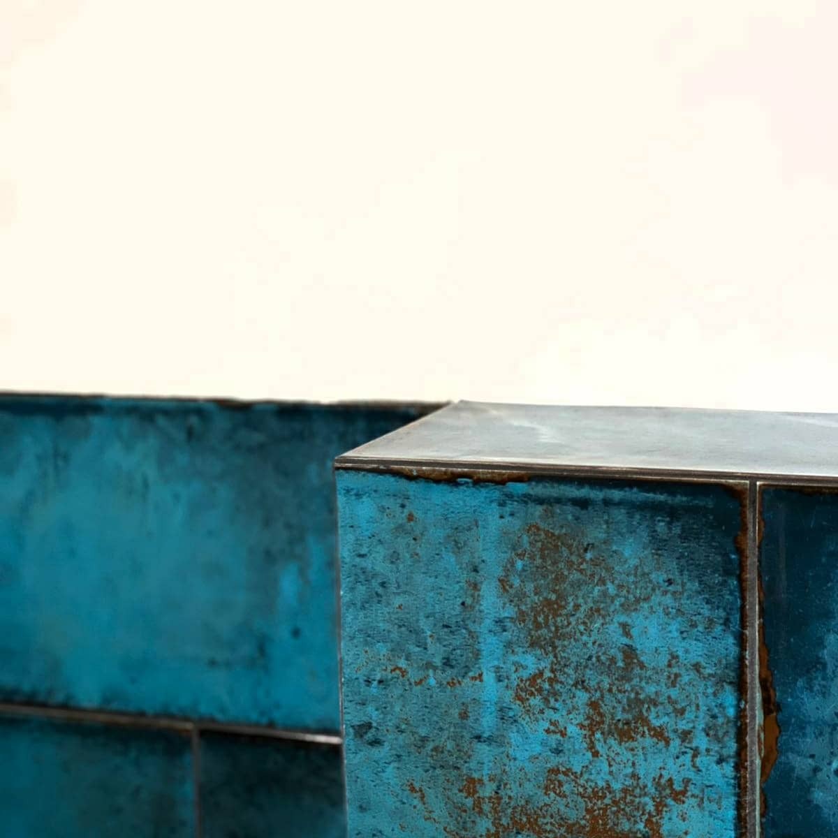 Narożne ujęcie ściany wyłożonej turkusowymi, metalizowanymi płytkami o rdzawym wzorze, z widoczną fugą i fragmentem białej ściany powyżej.