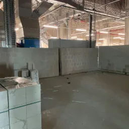 Wnętrze hali w trakcie budowy, widoczne ściany z bloczków, elementy instalacji pod sufitem oraz pracownik w odblaskowej kamizelce.