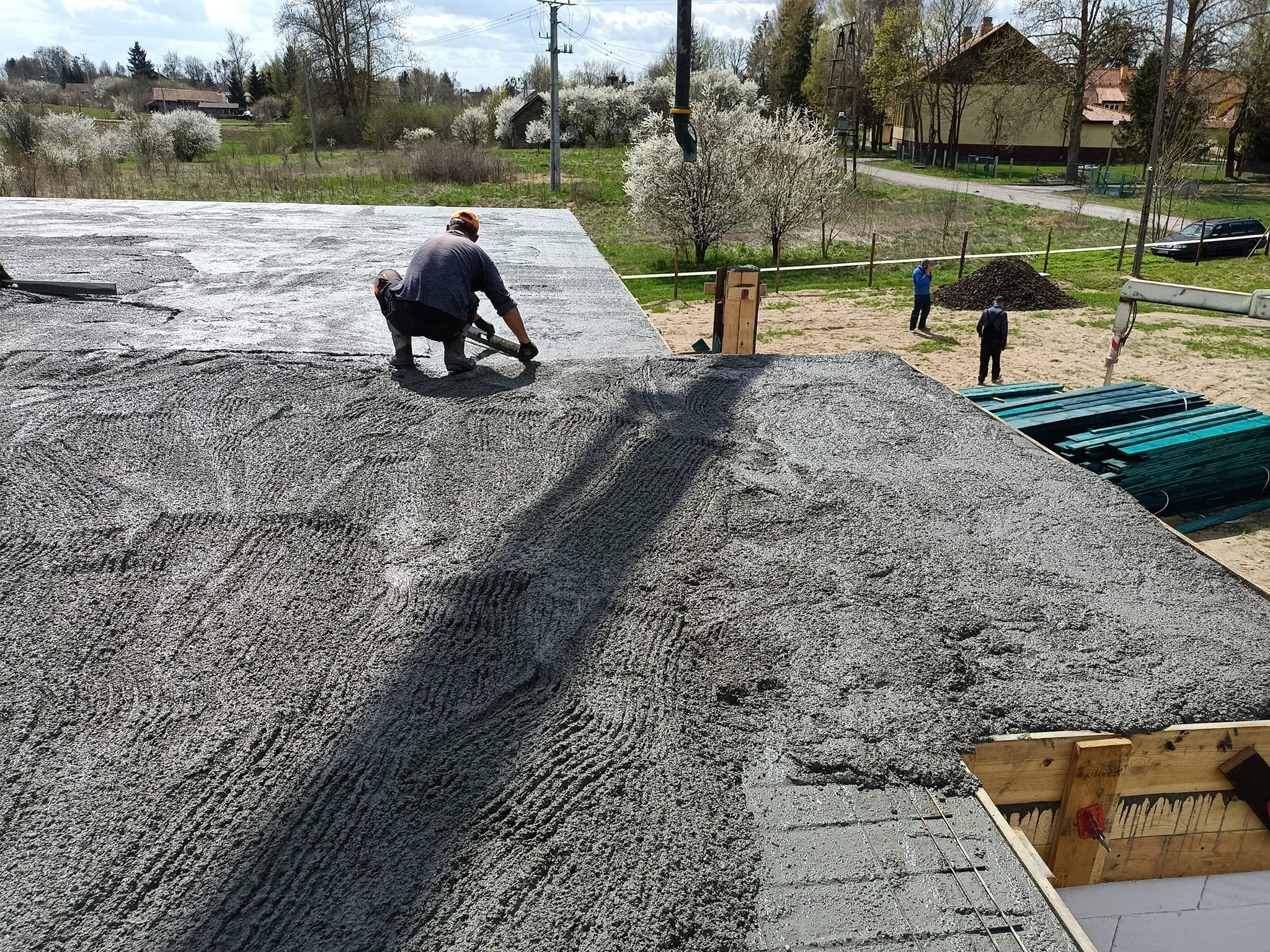 Pracownik budowlany wyrównuje świeżo wylany beton na płycie fundamentowej z widocznym zbrojeniem, w tle krajobraz wiejski z kwitnącymi drzewami.