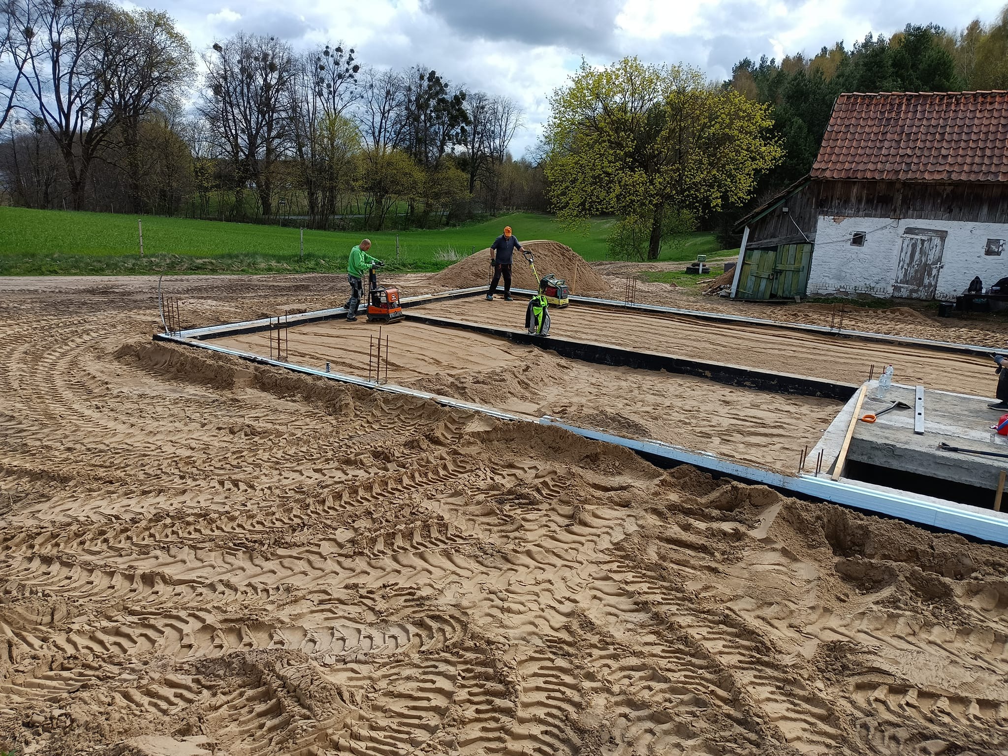 Przygotowanie terenu pod fundament: dwaj pracownicy zagęszczają piasek ubijarkami wibracyjnymi wewnątrz stalowego szalunku, w tle stary budynek gospodarczy i drzewa.