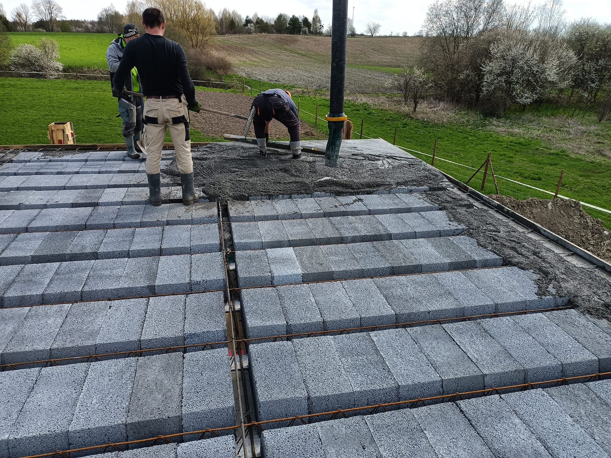 Wylewanie betonu na strop Teriva z widocznymi belkami i pustakami betonowymi, w tle pracownicy budowlani i krajobraz wiejski.