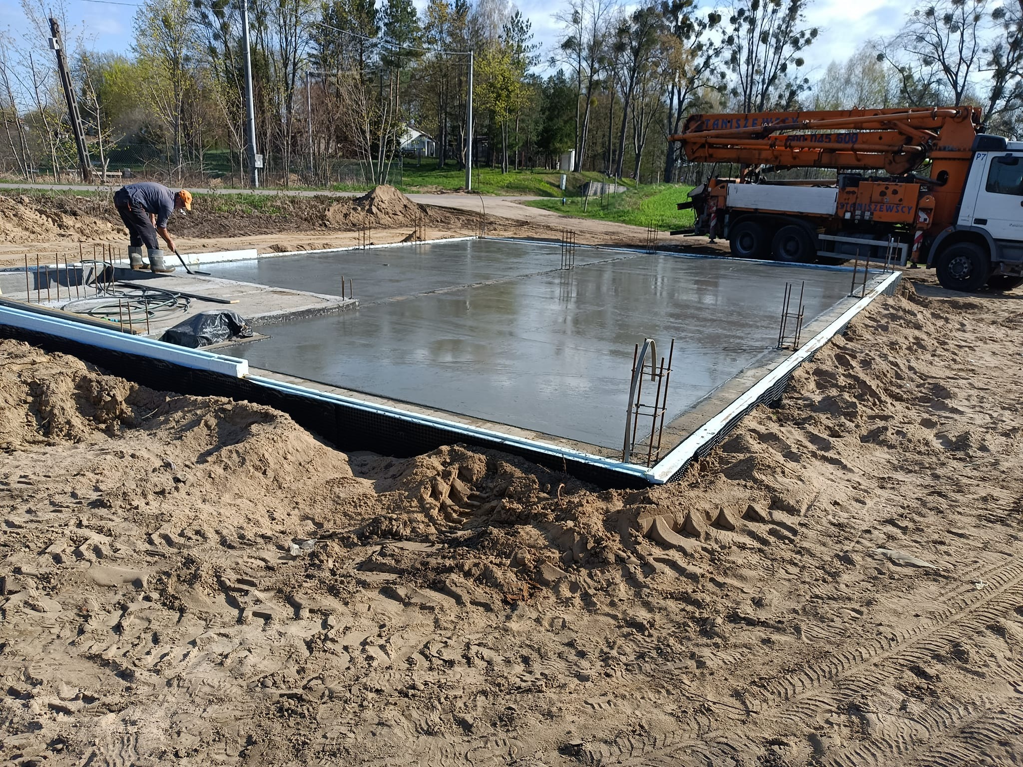 Wylewanie fundamentu domu jednorodzinnego: pracownik wygładza świeży beton na zbrojonej płycie fundamentowej, w tle pompa do betonu i drzewa.