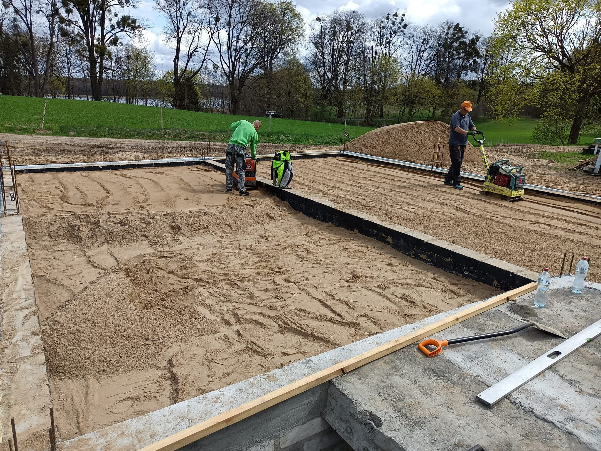 Przygotowanie podłoża pod płytę fundamentową: zagęszczanie piasku ubijarką wibracyjną, widoczna izolacja pionowa, szalunek, poziomica i dwie butelki wody.