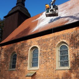 Renodach Serwis - Pracownik w pomarańczowej kamizelce na podnośniku koszowym czyści dach ceglanego kościoła z wieżą, strumieniem wody pod ciśnieniem.